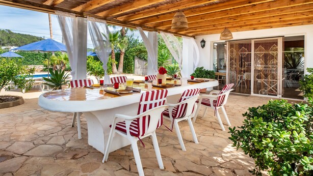 Un patio exterior con techo de madera, sillas blancas, una mesa redonda y sombrillas azules.