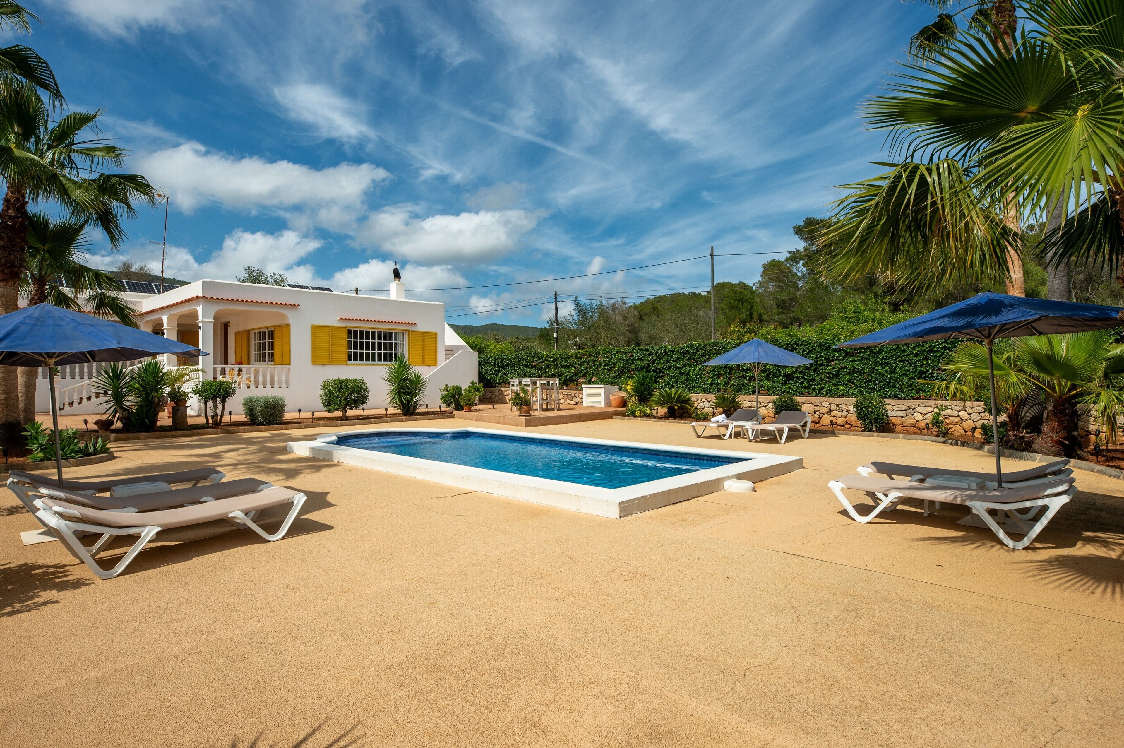 A pool with lounge chairs, a house with yellow shutters, and palm trees.