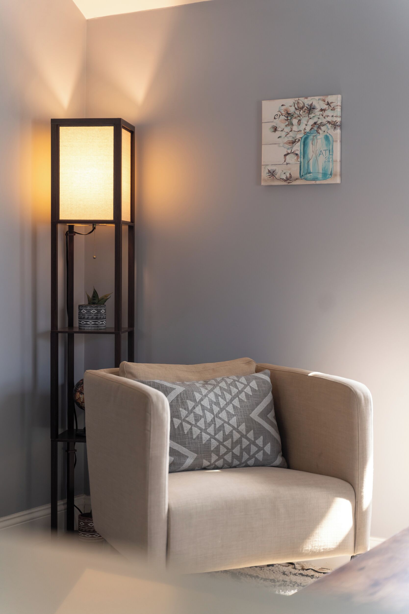 Ett hörnrum med en beige fåtölj, en golvlampa och en tavla som hänger på väggen.