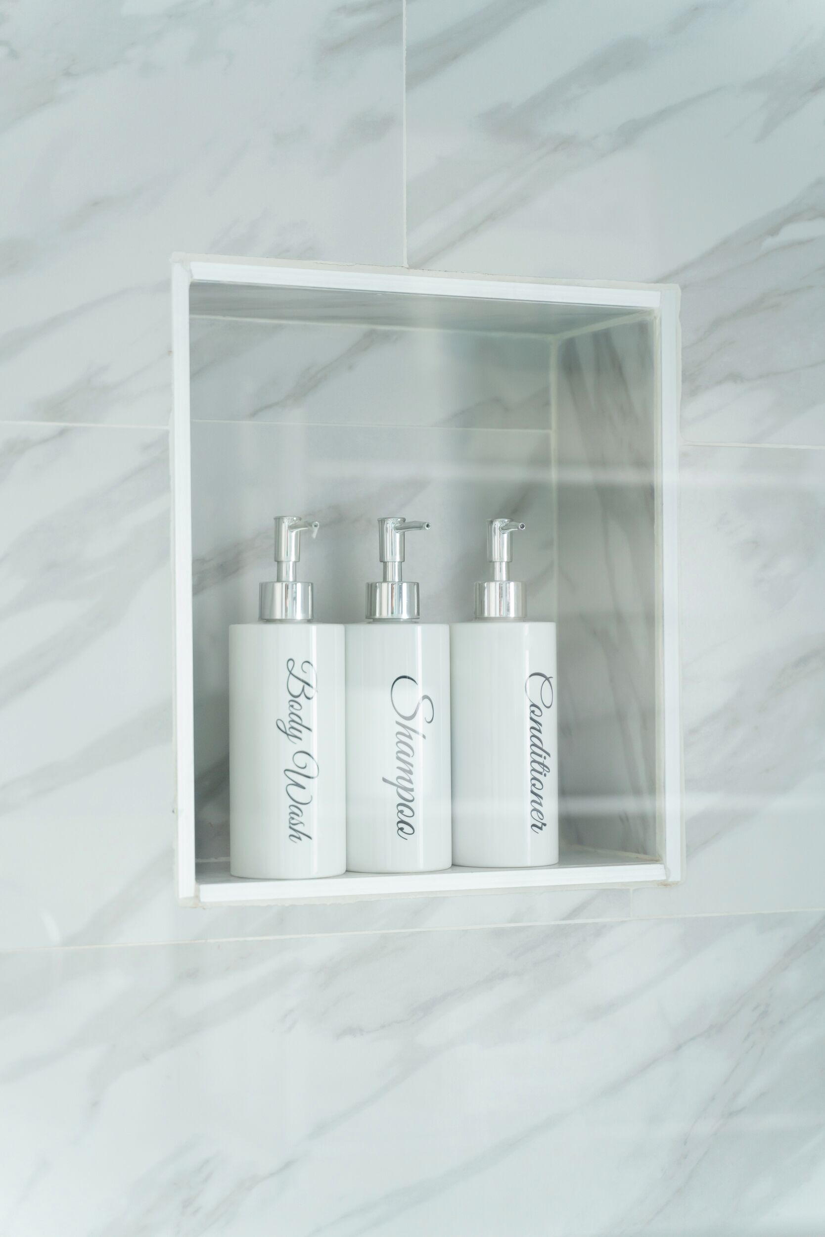A bathroom with a marble wall and a white shelf containing three bottles of body wash, shampoo, and conditioner.