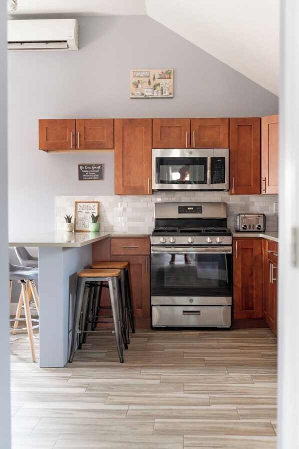 A modern kitchen with wooden cabinets, a stainless steel stove, a microwave, and a toaster. There are stools and a small island with a plant.