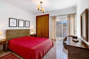 Une chambre d’hôtel avec un grand lit, une tête de lit en bois, un bureau avec un miroir, un balcon avec des rideaux et des tableaux encadrés au mur.