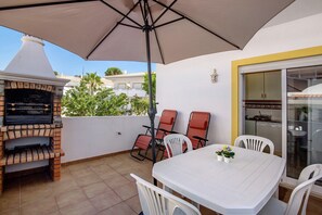 Una veranda con forno a mattoni, tavolo bianco, sedie e ombrellone.