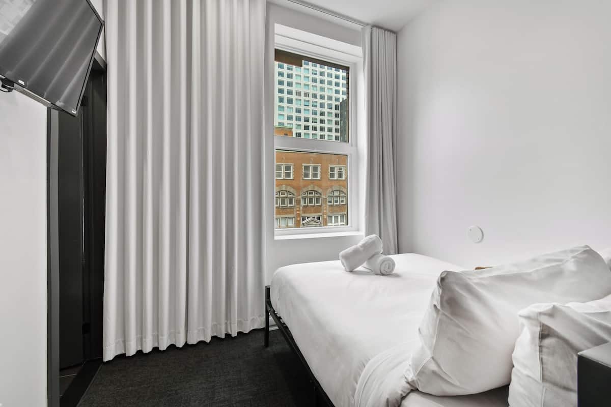 A hotel room with a bed, white curtains, a window with a view of buildings, and a flat-screen TV mounted on the wall.