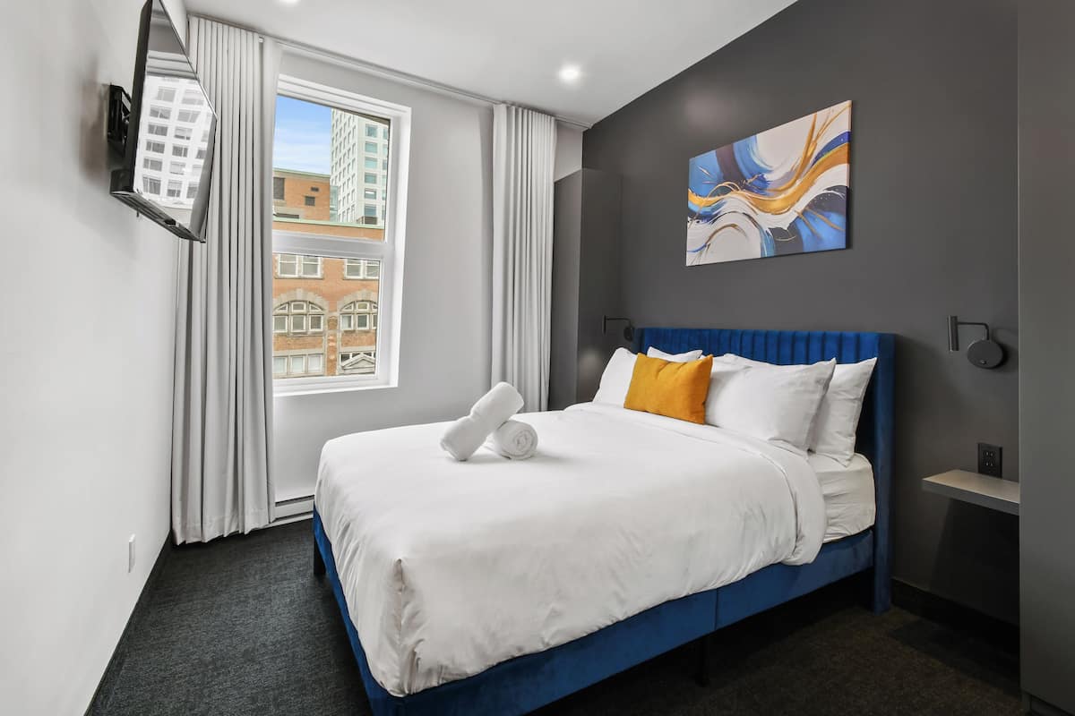 A hotel room with a large bed, a flat-screen TV mounted on the wall, a window with curtains, and a city view.