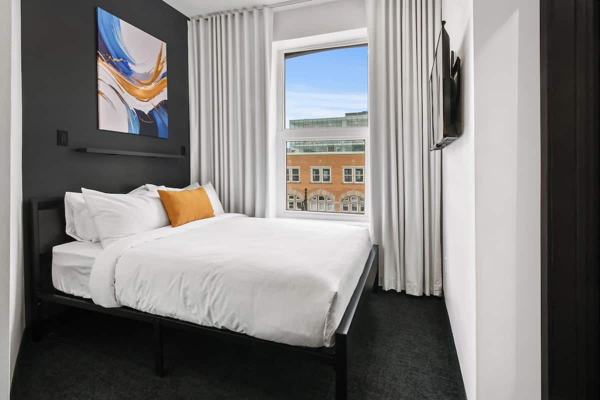 A modern hotel room with a large bed, a view of a building through a window, and a wall-mounted TV.