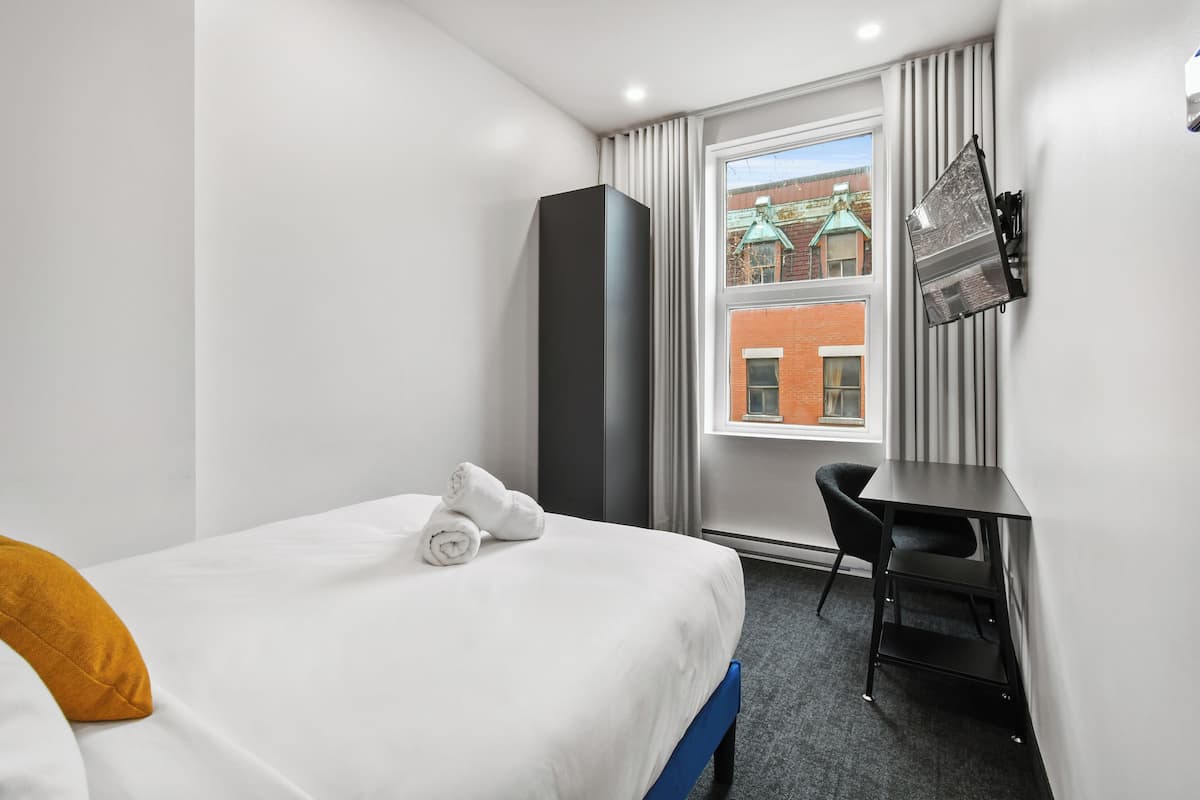 A hotel room with a bed, a desk, a chair, a window with a view of buildings, and a wall-mounted TV.