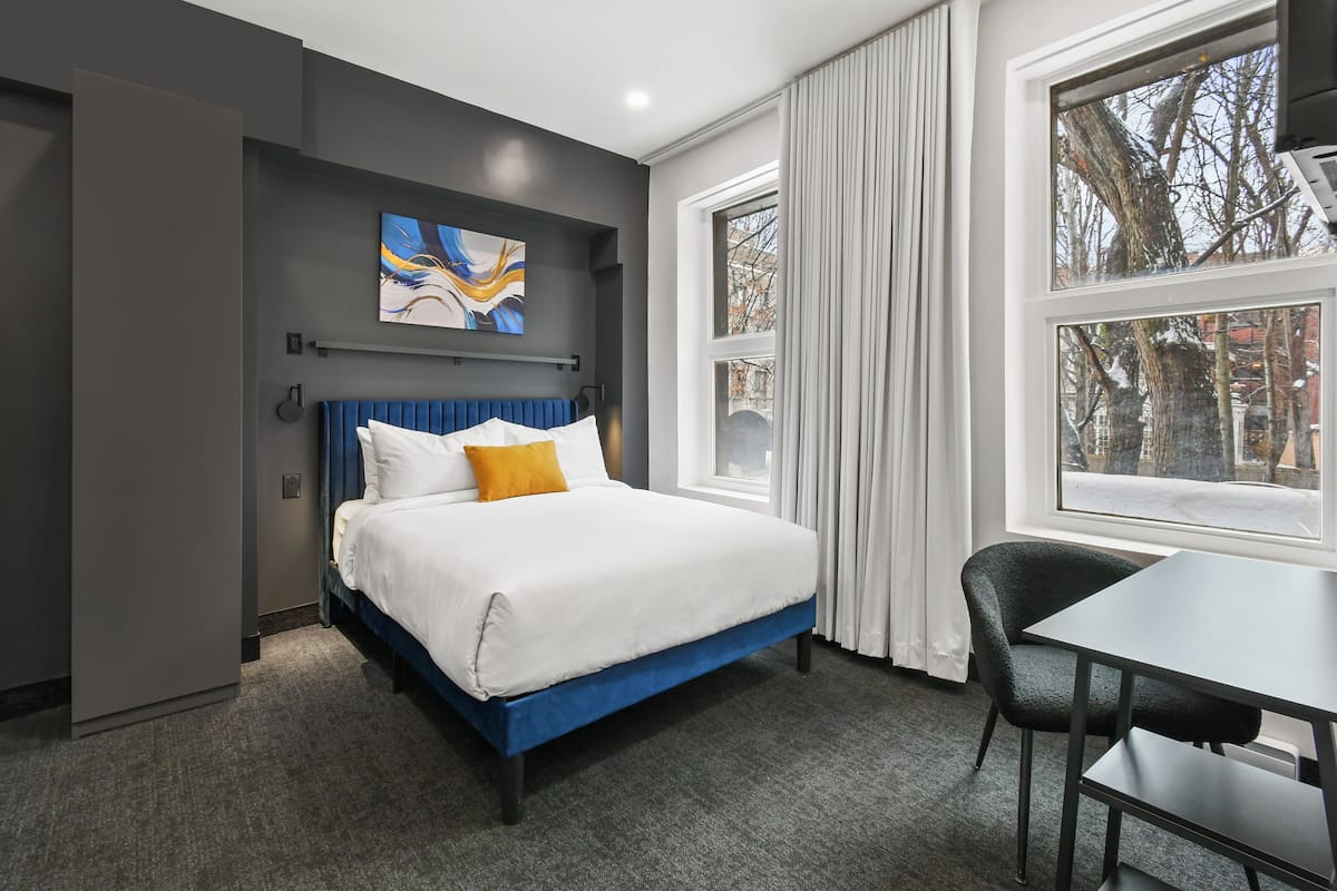 A modern hotel room with a blue bed, white bedding, a gray headboard, and a large window with curtains.