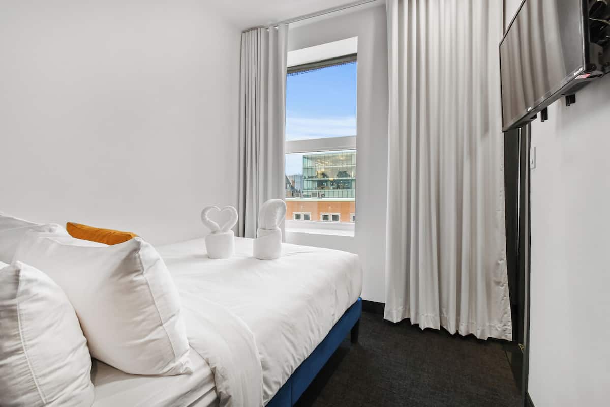 A hotel room with a bed, white curtains, a window with a view of buildings, and a blue headboard.