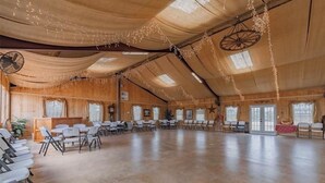 A large hall with a high ceiling, wooden walls, and a fan. Tables and chairs are arranged for an event.