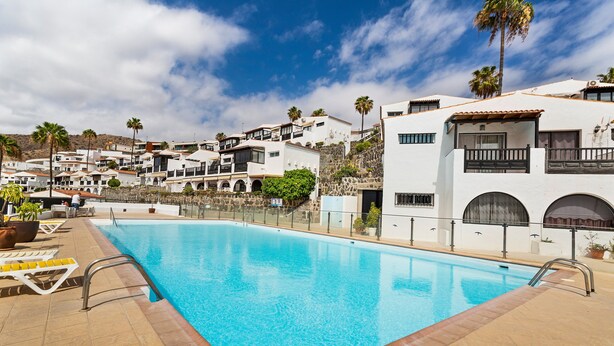 A swimming pool with lounge chairs, a stone wall, and white buildings with balconies. - Holiday Apartment 'Bungalow Josephin' with Shared Pool, Private Terrace and Wi-Fi (Patalavaca)