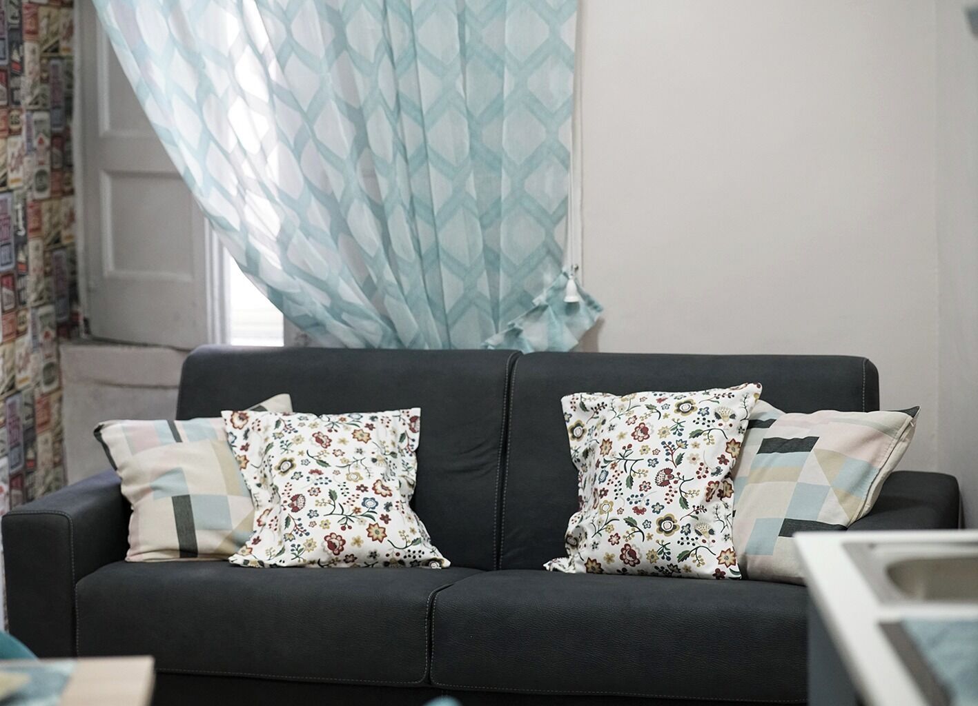 A black sofa with floral-patterned cushions and a patterned curtain in a room with a plain wall and a window.