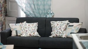 A black sofa with floral-patterned cushions and a patterned curtain in a room with a plain wall and a window.