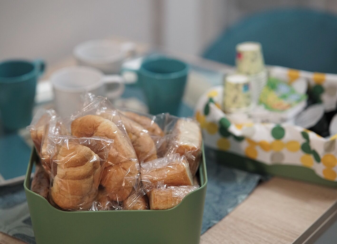 Uma caixa verde cheia de pães, um tabuleiro com padrão de bolinhas amarelas e brancas, e vários copos cor de turquesa sobre uma mesa.