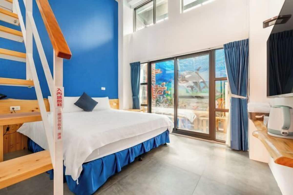 A hotel room with a bed, a wooden ladder, a large window with curtains, and a view of the outside.