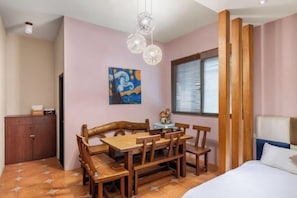 A room with a wooden dining table and chairs, a bed with white bedding, a painting on the wall, and a window with blinds. - Xinhaiminsu (Hualien)