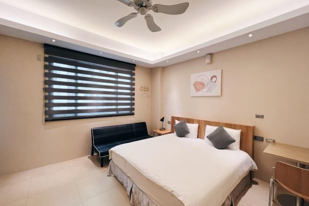 A modern bedroom with a large bed, a ceiling fan, a bench, a bedside table, and a wall-mounted artwork.