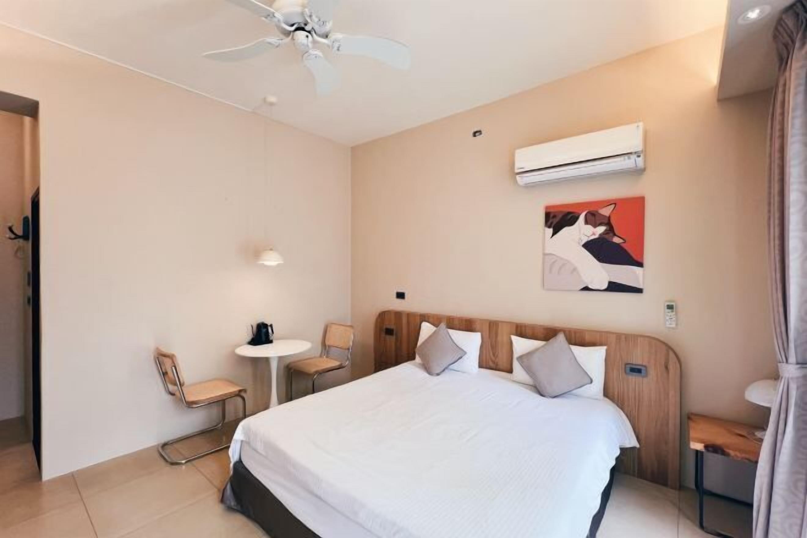 Une chambre d’hôtel avec un lit, un ventilateur de plafond, une petite table avec une chaise, un appareil de climatisation et un tableau au mur.