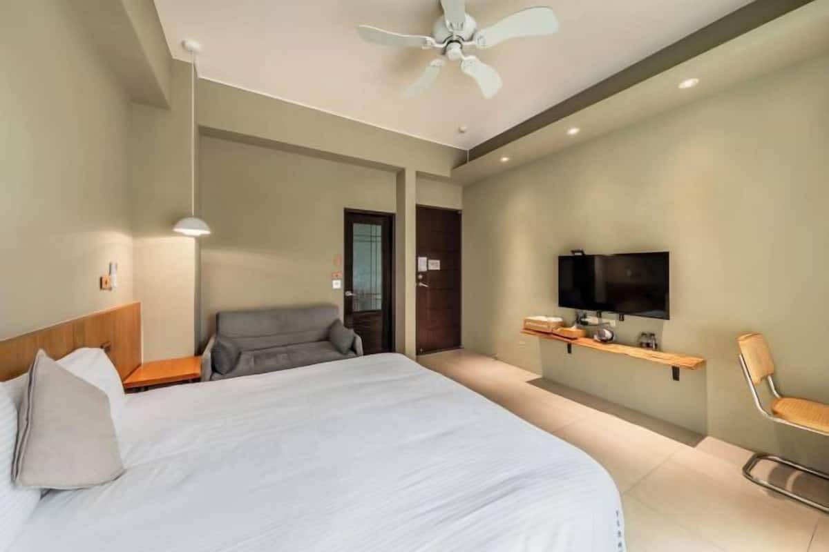 A modern hotel room with a large bed, a ceiling fan, a TV, and a seating area with a chair.