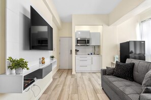 A modern living room with a flat-screen TV, grey sofa, and a small kitchen area with white cabinets and a microwave.