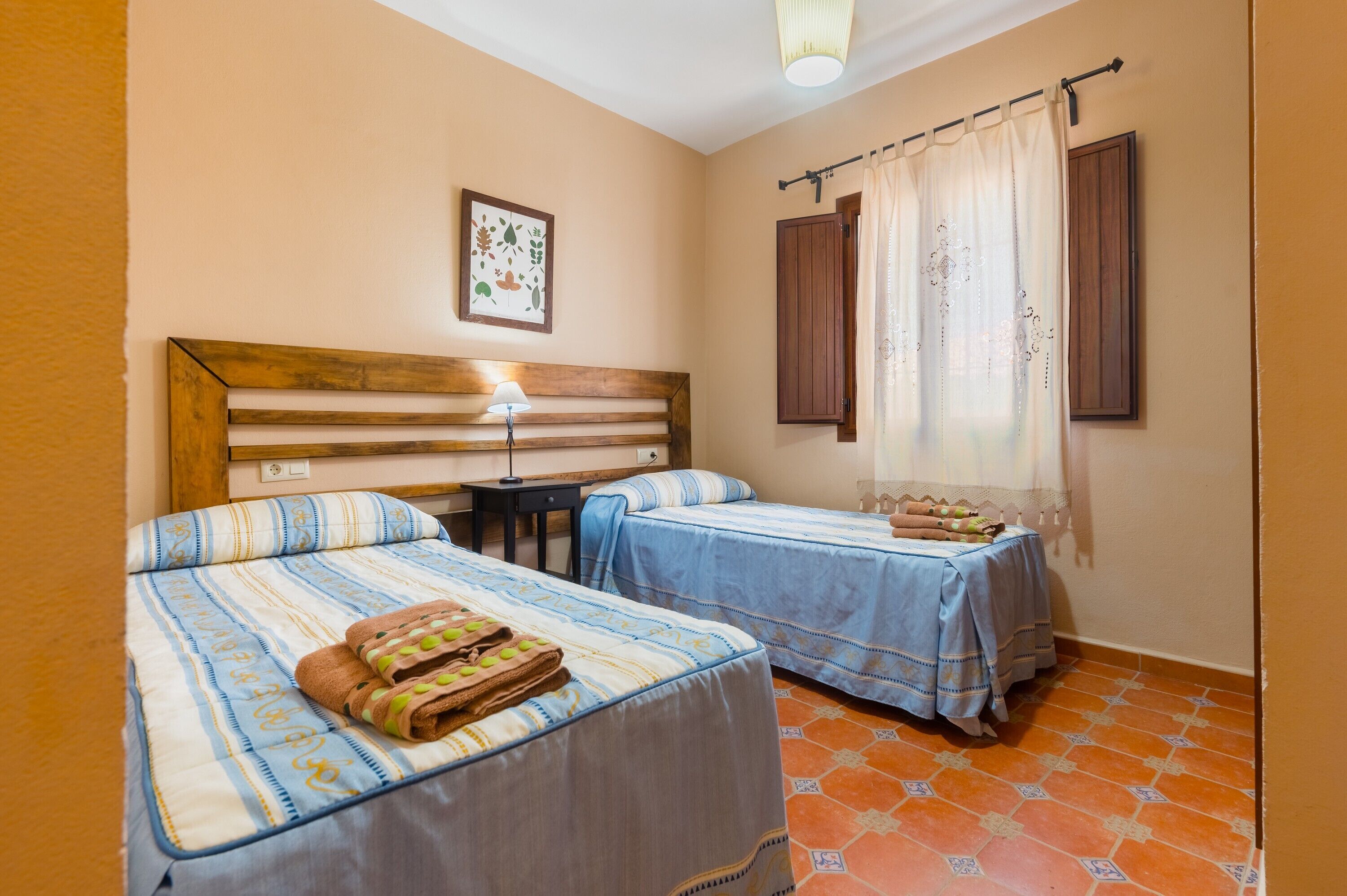 A room with two beds, a wooden headboard, a bedside table with a lamp, a framed picture on the wall, and a window with curtains.