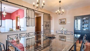 Zona pranzo con tavolo in vetro, sedie di vimini, un lampadario e un pensile della cucina sullo sfondo.