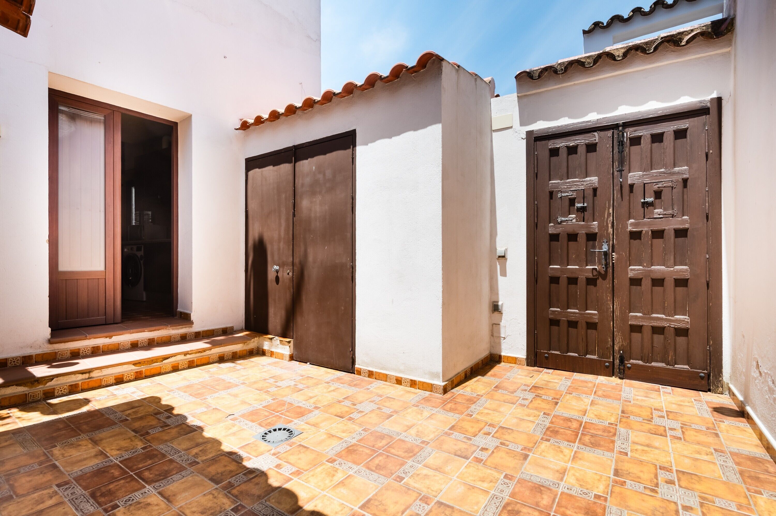 Un patio con suelo de baldosas, una puerta de madera y una pared blanca.