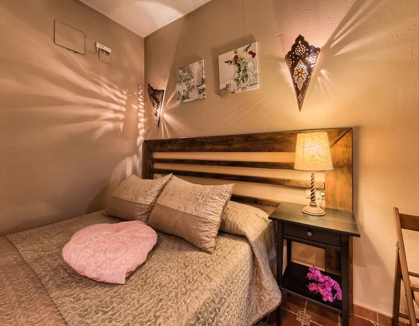 Une chambre à coucher avec un lit, des oreillers, une table de chevet, une lampe, des œuvres d’art murales et une tête de lit en bois.