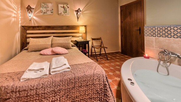 A bedroom with a large bed, a wooden headboard, a bedside table with a lamp, a chair, a bathroom with a bathtub, and a door.