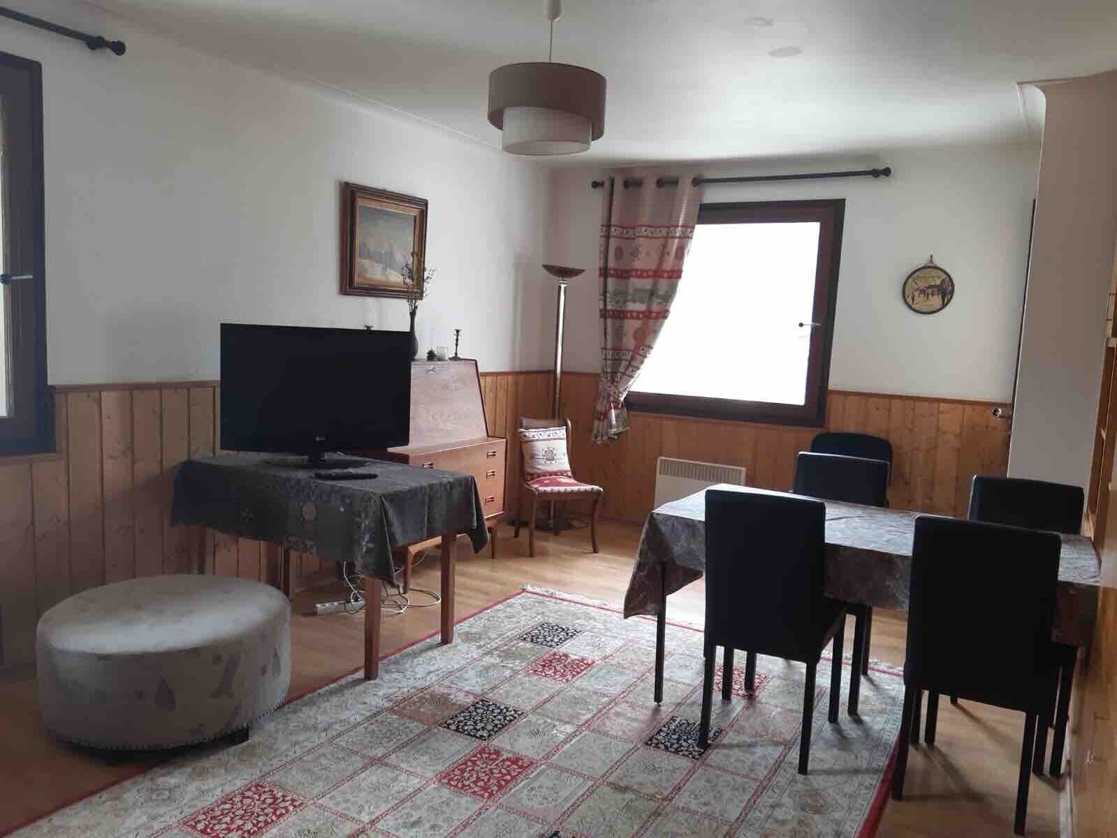 A dining area with a table and chairs, a television on a stand, a patterned rug, and a window with curtains.