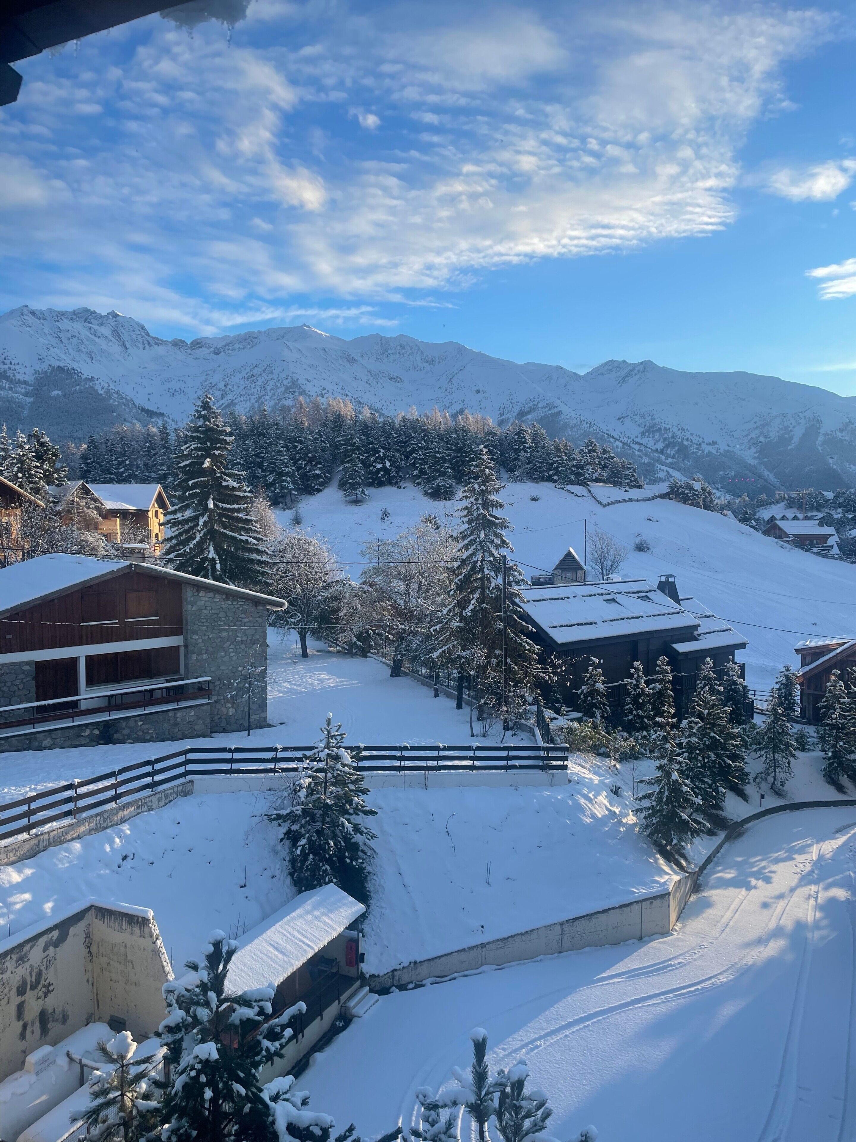 Un village de montagne enneigé, avec des bâtiments traditionnels en bois, une structure en pierre et une vue sur les montagnes en arrière-plan.