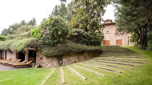A stone building with a staircase leading up to it, surrounded by lush greenery and a grassy area with wooden lounge chairs. - Country Home 'Gran Castanyer' with Mountain View, Private Pool and Wi-Fi (Viladrau)