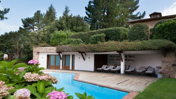 A poolside area with a wooden deck, lounge chairs, and a stone building with a green roof. - Country Home 'Gran Castanyer' with Mountain View, Private Pool and Wi-Fi (Viladrau)