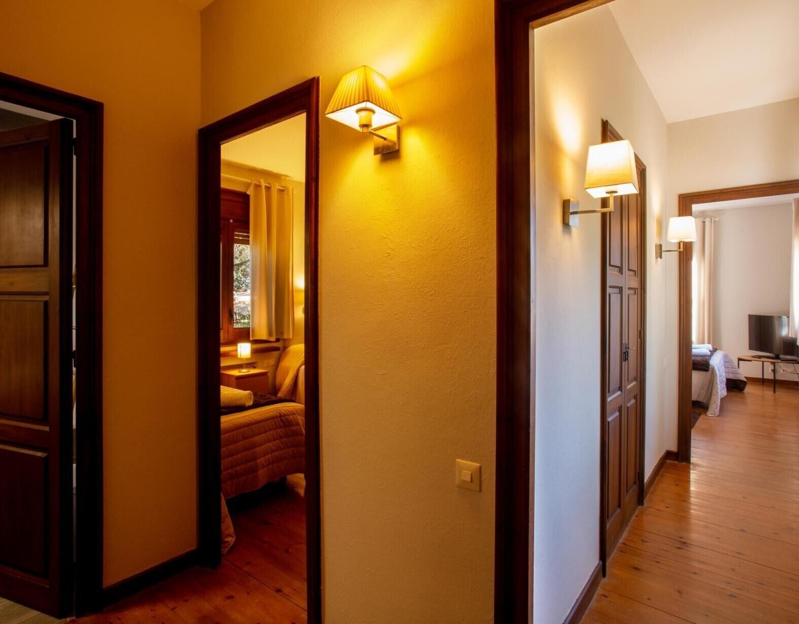 Un couloir d’hôtel avec un plancher en bois, un lit en arrière-plan et des lampes fixées au mur.