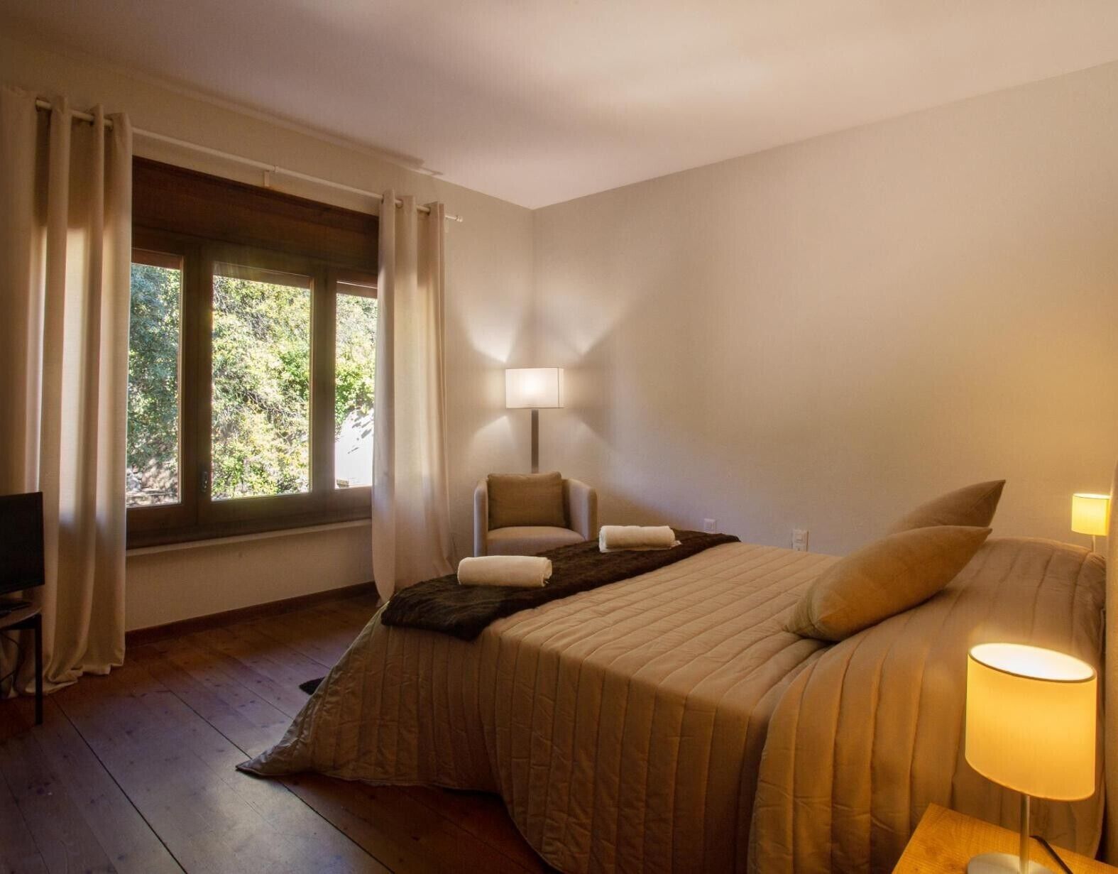 Habitació d'hotel amb llit, cadira, taula, làmpada i finestra amb cortines.