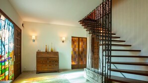 Una escalera con barandilla de metal, un aparador de madera, una puerta de vitral y un aplique de pared.