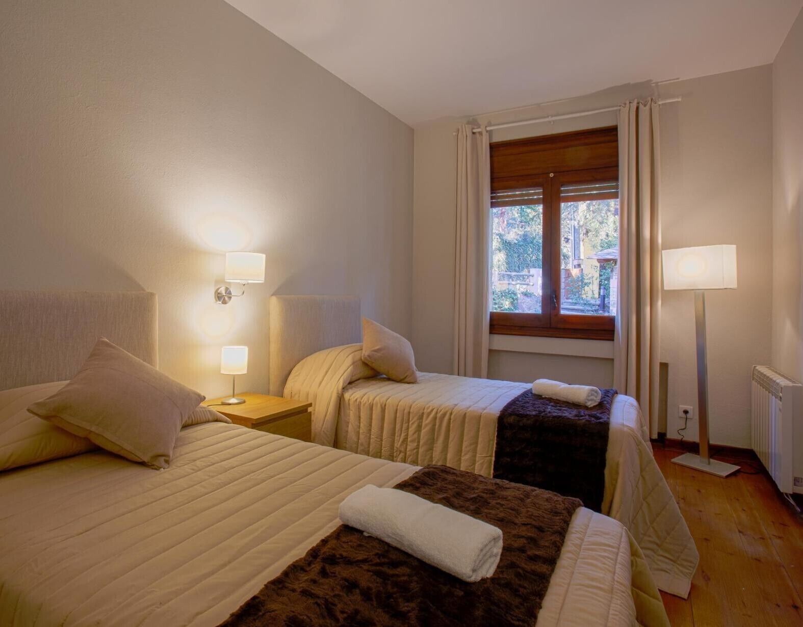 Habitación de hotel con dos camas, una ventana con cortinas, una mesita de noche con lámpara y un radiador.