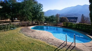 Una piscina con solárium de piedra, rodeada de césped y árboles, y montañas al fondo.