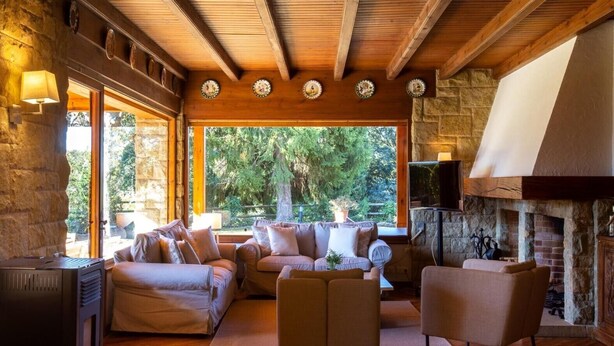 Una sala de estar acogedora con chimenea de piedra, vigas de madera y un amplio ventanal con vistas al verdor.