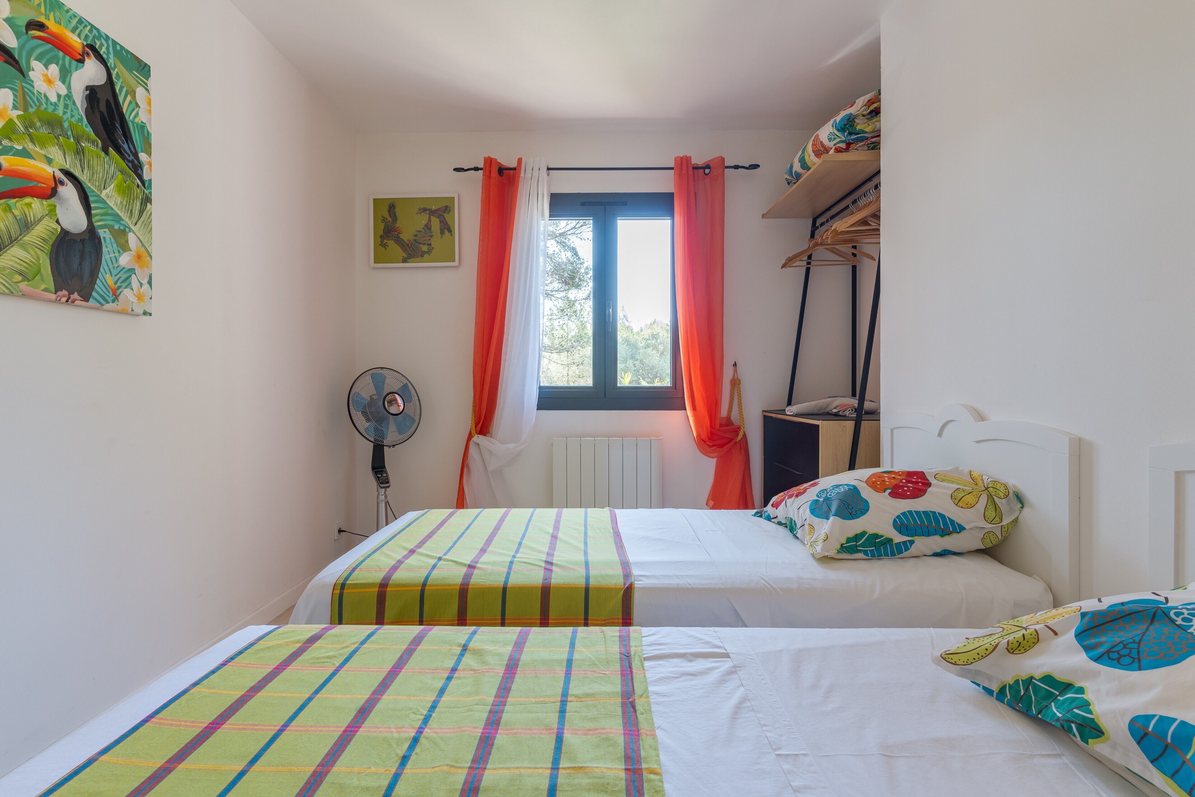 A bedroom with two beds, a window with curtains, a radiator, a fan, and a wall with a tropical bird painting.