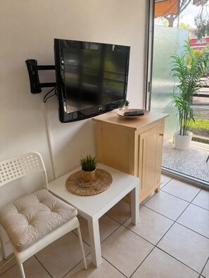 A room with a mounted TV, a small wooden cabinet, a white chair, and a potted plant on a white table. - 06U - Beautiful studio in residence - swimming pool and tennis court (Saint-Laurent-du-Var)