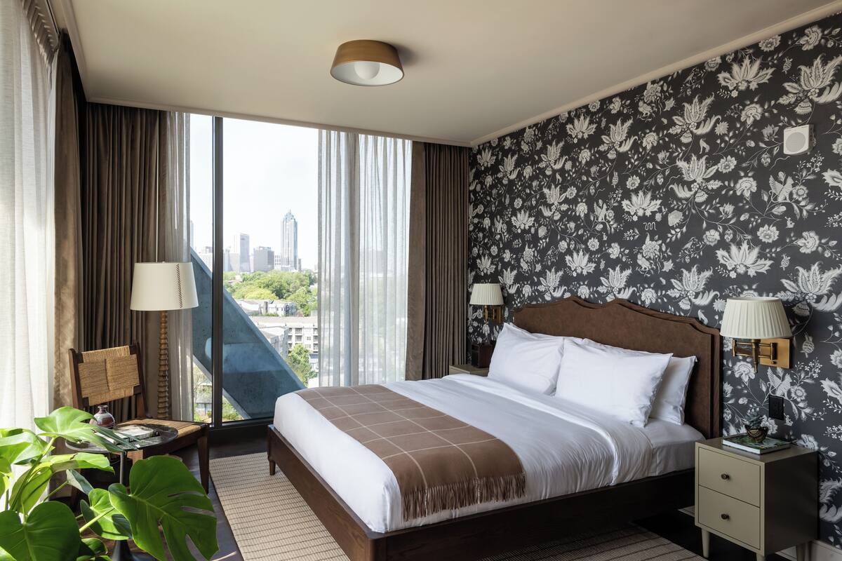 A hotel room with a large bed, two bedside lamps, a view of the city, and a floral wallpaper.