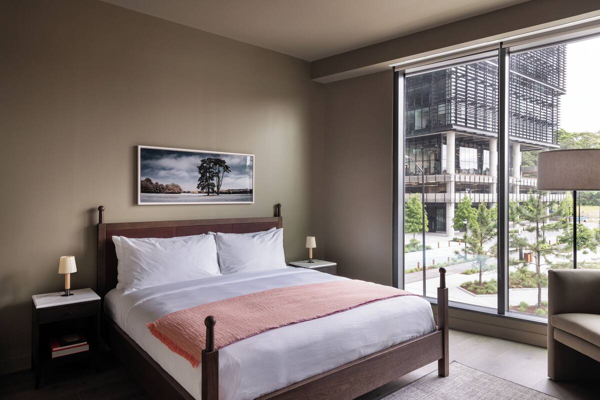 A bedroom with a large window, a bed, a nightstand, a chair, and a painting on the wall.