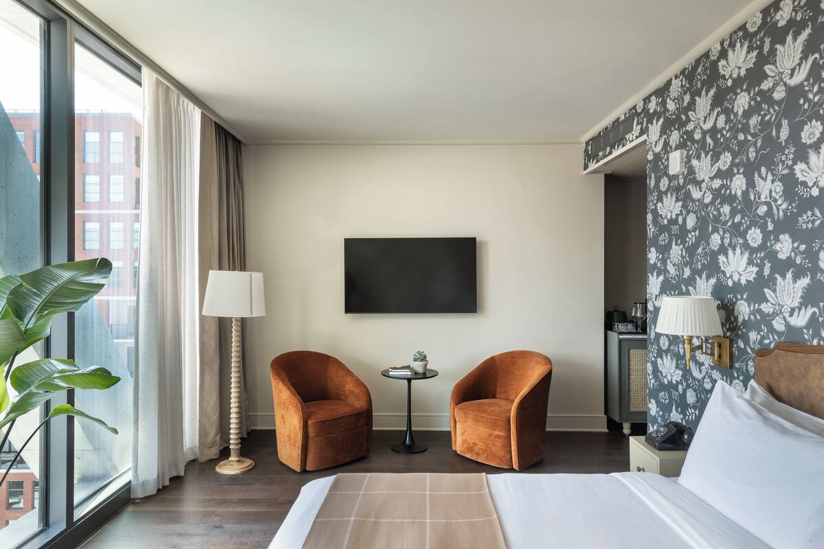 A hotel room with a bed, two armchairs, a small table, a TV, and a large window with curtains.