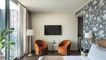 Une chambre d’hôtel avec un lit, deux fauteuils, une petite table, un téléviseur et une grande fenêtre avec des rideaux.