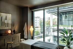 A modern hotel room with a large window, a bed, a bedside table with a lamp, a plant, and a view of the cityscape.