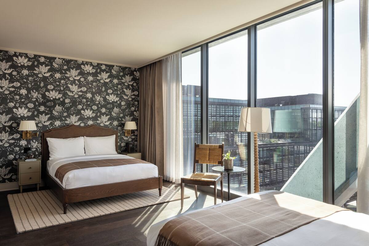 A modern hotel room with a large bed, a small table with a chair, and a view of the city through large windows.