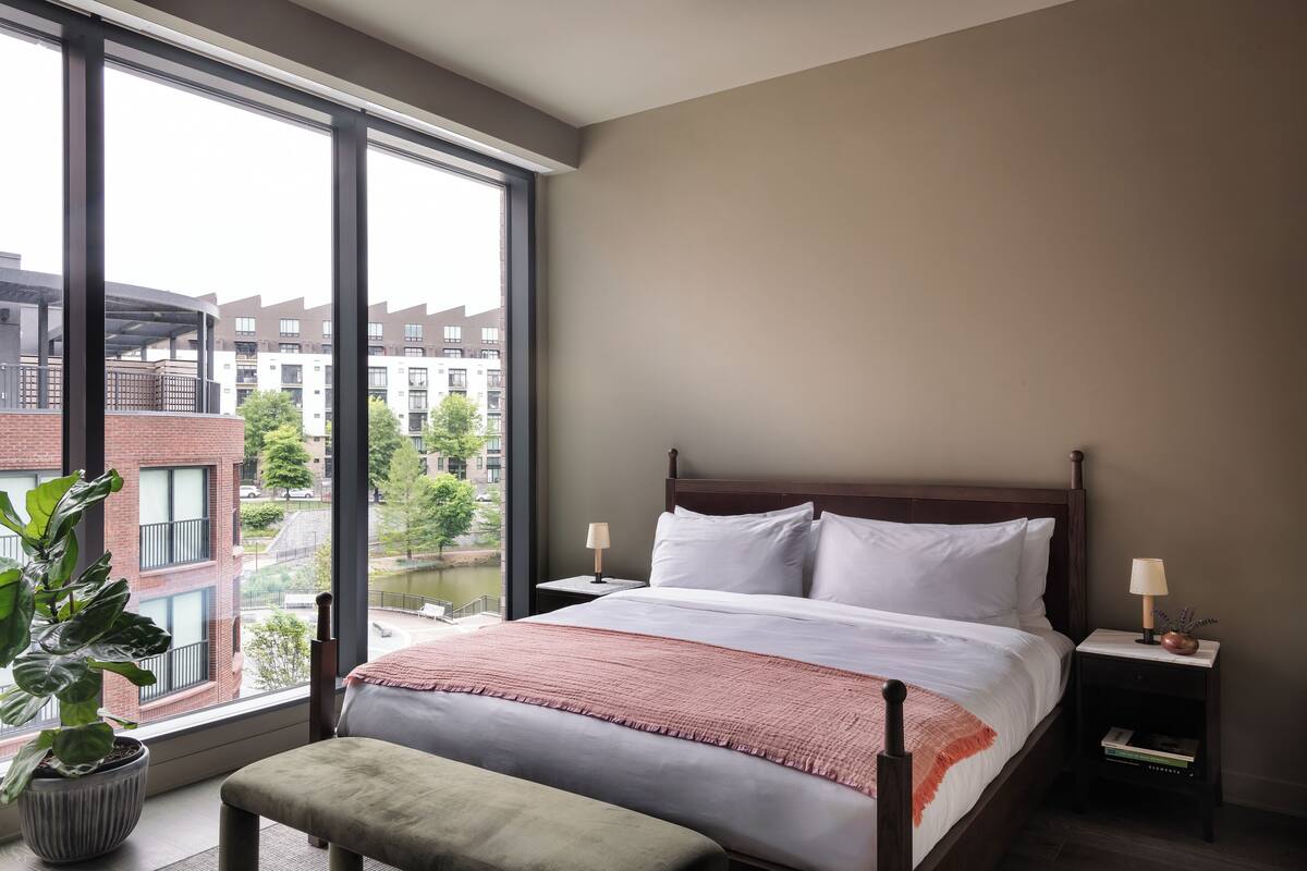 A bedroom with a large window, a bed, a nightstand, a lamp, and a potted plant.