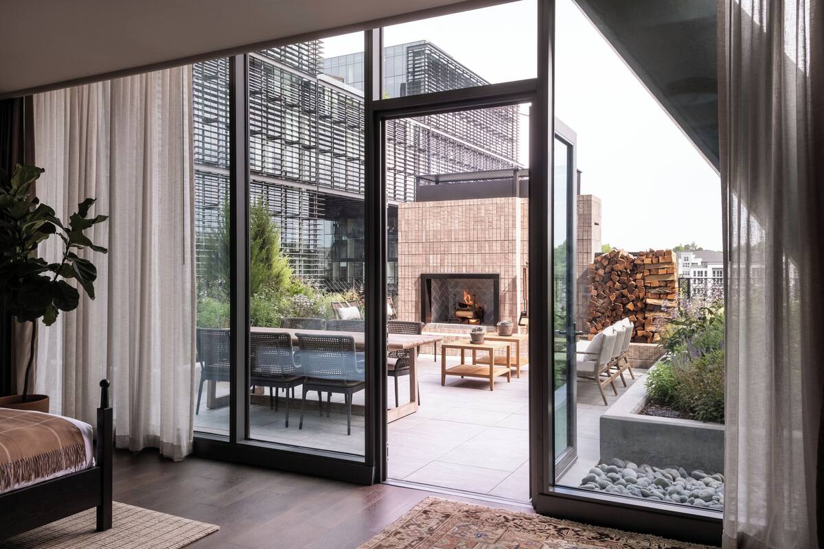 A modern room with a large sliding glass door leading to an outdoor patio with a fireplace, seating, and a city view.