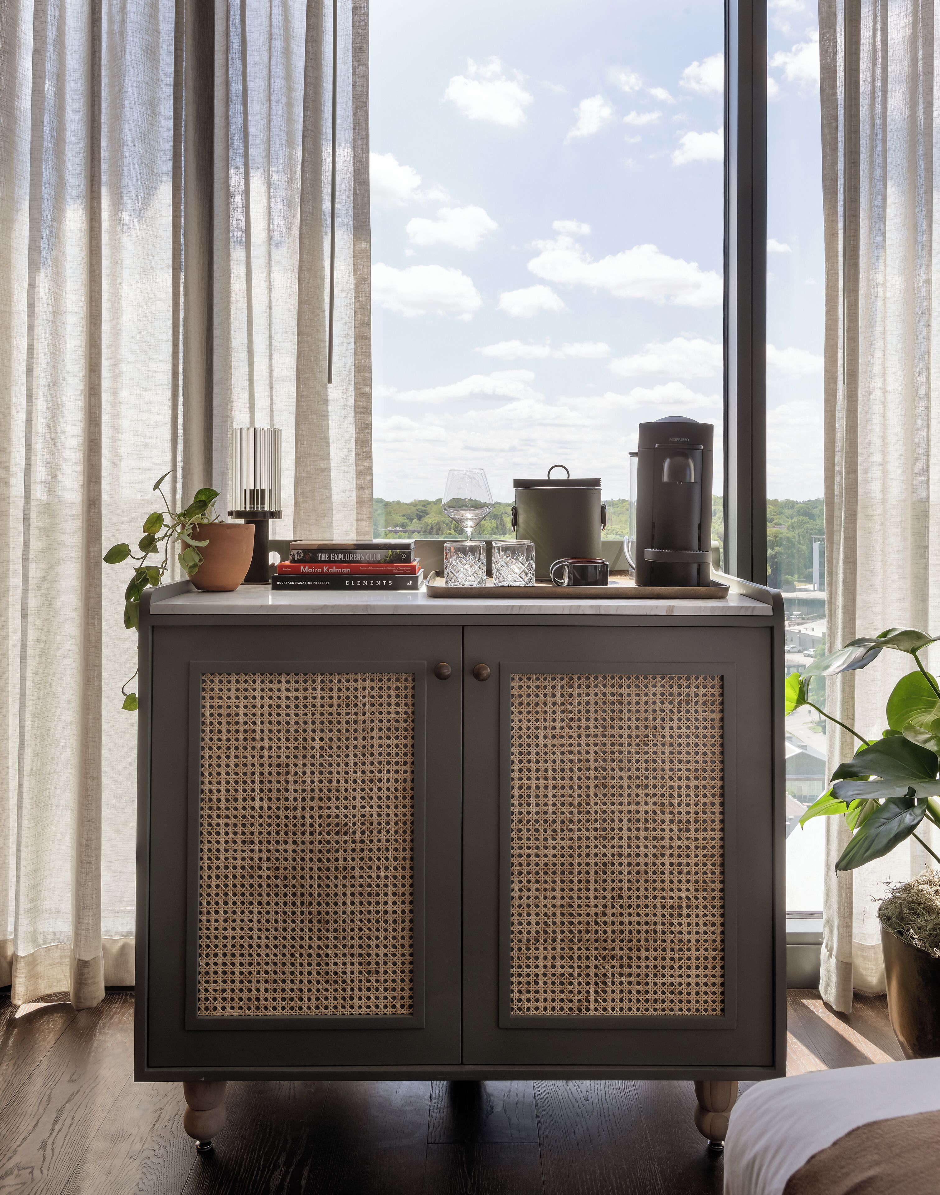 Ein Holzschrank mit Flechtpaneelen, ein Topfpflanze, ein Buch, ein Glas, ein Wasserkocher und eine Kaffeemaschine auf einem Fensterbrett.